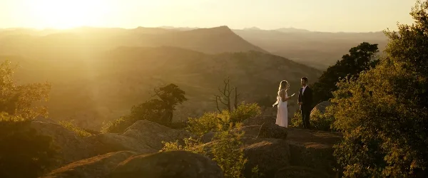 A bride and groom standing on a rocky mountain overlook during a golden hour sunset.