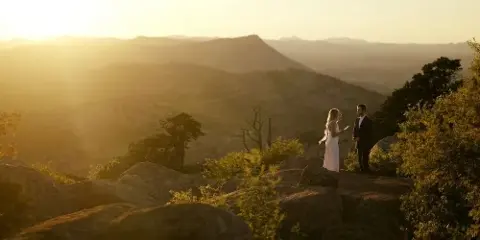 A bride and groom standing on a rocky mountain overlook during a golden hour sunset.