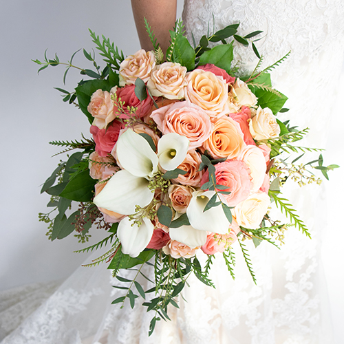 A hand holding a lush wedding bouquet composed of peach roses, white calla lilies, and greenery.