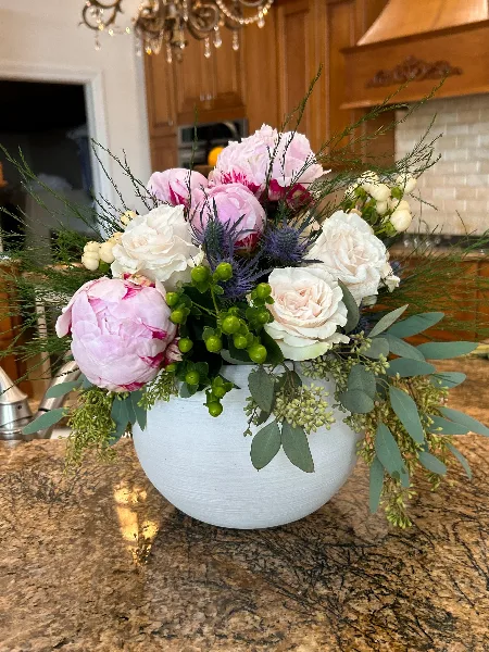 A lush floral arrangement with pink peonies, white roses, and greenery in a white vase on a kitchen counter.
