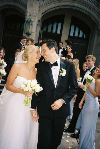 A bride and groom smiling and holding hands with their wedding party in the background.