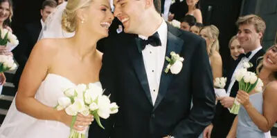A bride and groom smiling and holding hands with their wedding party in the background.