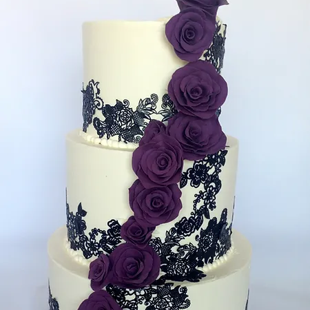 A three-tier white wedding cake decorated with black lace-style modeling chocolate and a cascading arrangement of purple roses.