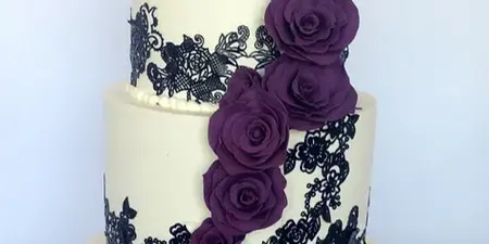 A three-tier white wedding cake decorated with black lace-style modeling chocolate and a cascading arrangement of purple roses.