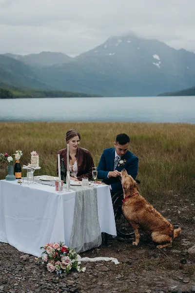 A bride and groom seated at a decorated outdoor wedding table with a dog, set against a backdrop of a lake and mountains.