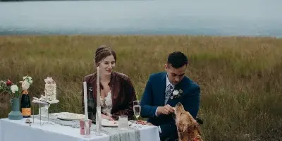 A bride and groom seated at a decorated outdoor wedding table with a dog, set against a backdrop of a lake and mountains.