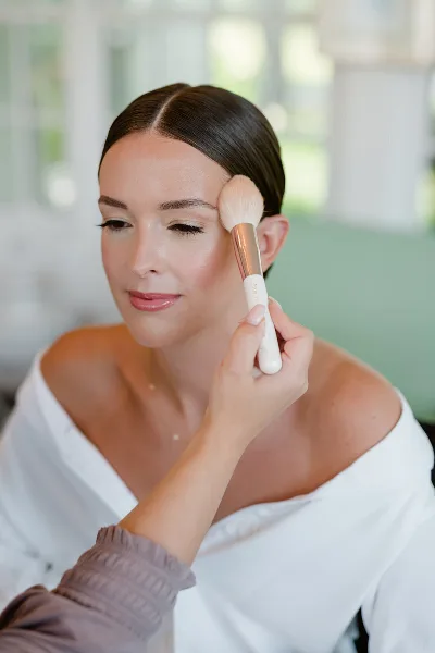 A makeup artist applying powder to a bride's cheek with a brush.