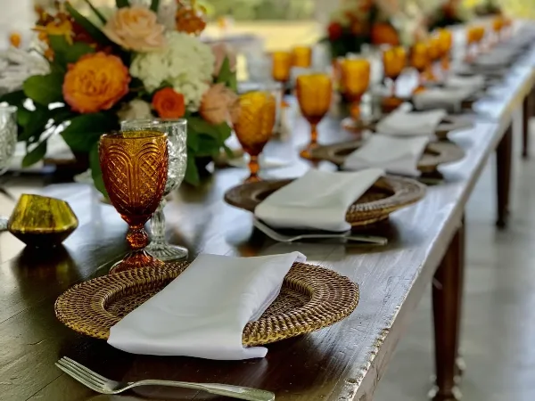 Long event table set with woven chargers, white napkins, amber glassware, and floral centerpieces.