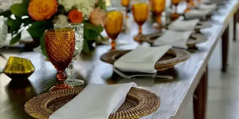 Long event table set with woven chargers, white napkins, amber glassware, and floral centerpieces.