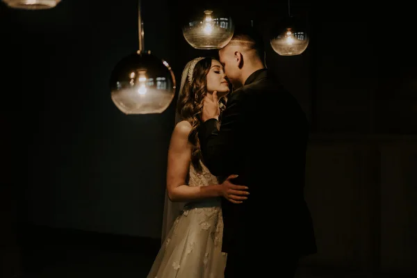 A bride and groom sharing a kiss under hanging glass globe lights in a dimly lit setting.