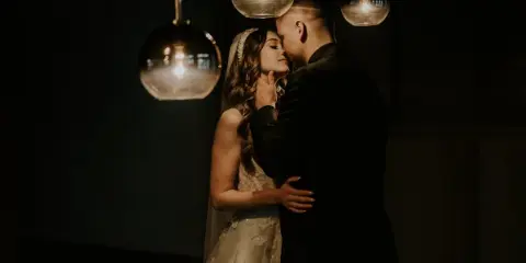 A bride and groom sharing a kiss under hanging glass globe lights in a dimly lit setting.