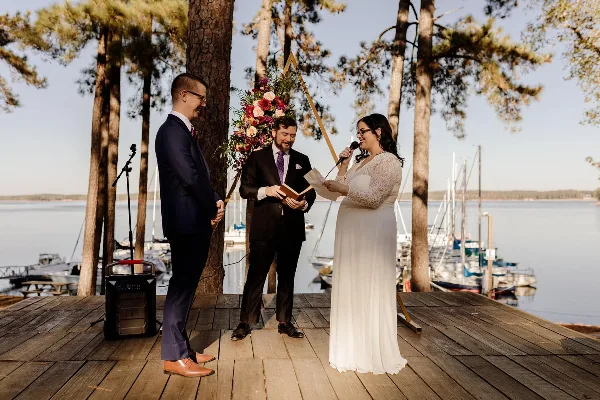 A wedding ceremony taking place on a wooden dock by a lake, featuring a bride, groom, and officiant.