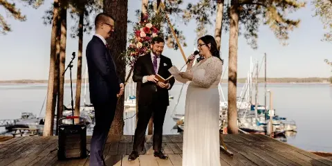 A wedding ceremony taking place on a wooden dock by a lake, featuring a bride, groom, and officiant.