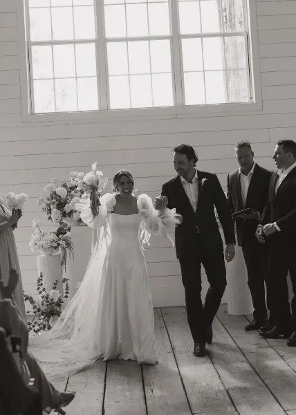 A bride and groom walking down the aisle during a wedding ceremony in a white-walled venue with floral arrangements.