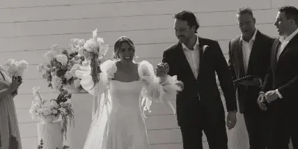 A bride and groom walking down the aisle during a wedding ceremony in a white-walled venue with floral arrangements.
