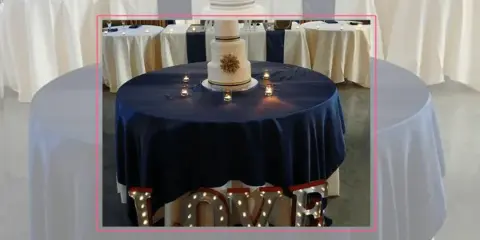 A wedding cake table decorated with a navy blue tablecloth, candles, and illuminated LOVE letters.