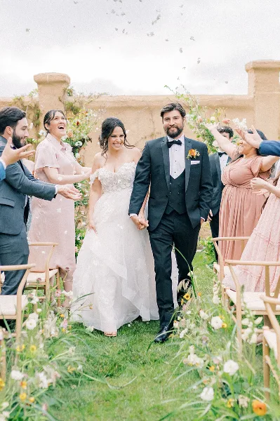 A bride and groom walking down a floral-lined aisle, smiling and holding hands as guests throw petals.