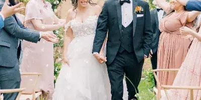 A bride and groom walking down a floral-lined aisle, smiling and holding hands as guests throw petals.