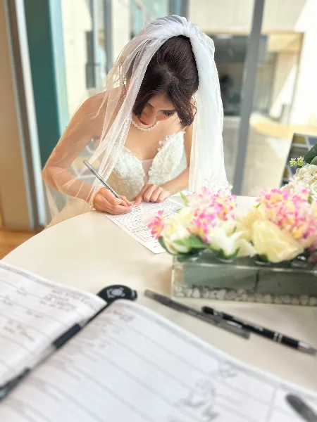 A bride in a white wedding dress and veil signing a marriage license document at a table with floral arrangements.