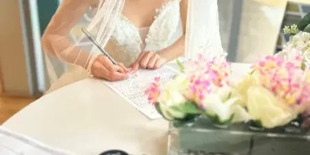 A bride in a white wedding dress and veil signing a marriage license document at a table with floral arrangements.