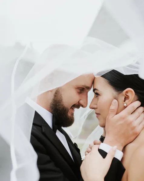 A bride and groom looking at each other under a veil, with the bride's hair and makeup visible.