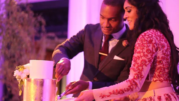 A bride in a red-and-white embroidered gown and a groom in a gray suit cutting their wedding cake together under purple and pink reception lighting.