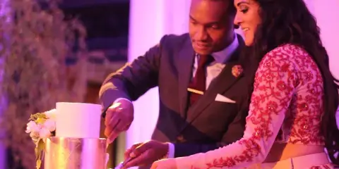 A bride in a red-and-white embroidered gown and a groom in a gray suit cutting their wedding cake together under purple and pink reception lighting.