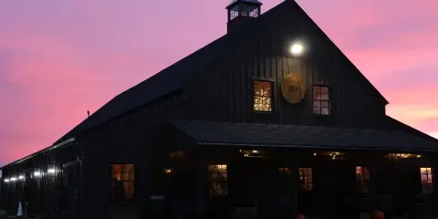 Exterior view of the SaltWater Fields event barn at dusk, featuring a dark wooden facade, a cupola, and a circular logo on the wall.