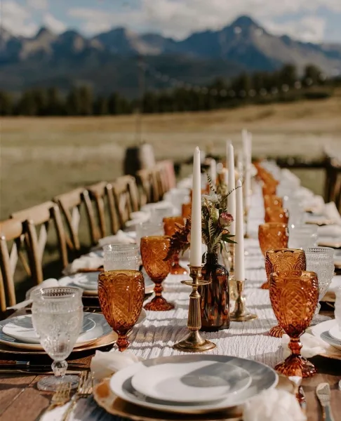An elegant outdoor dining table set with amber glassware, white plates, and tall candles against a mountain backdrop.