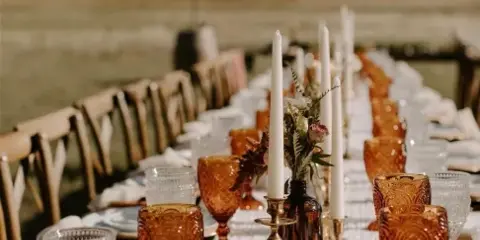 An elegant outdoor dining table set with amber glassware, white plates, and tall candles against a mountain backdrop.