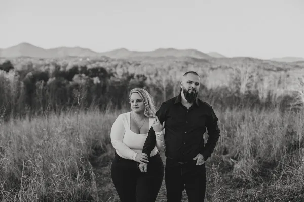 Black and white outdoor portrait of a couple standing together in a grassy field with mountains in the background.