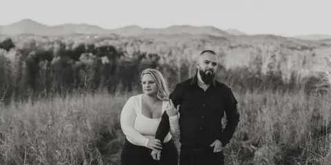 Black and white outdoor portrait of a couple standing together in a grassy field with mountains in the background.
