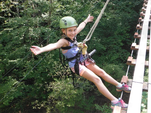 A young girl wearing a helmet and safety harness smiles while riding a zip line through a forest.