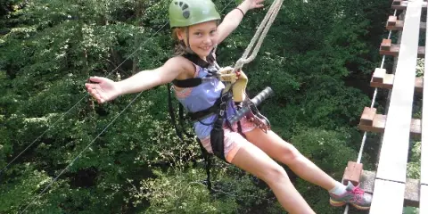 A young girl wearing a helmet and safety harness smiles while riding a zip line through a forest.