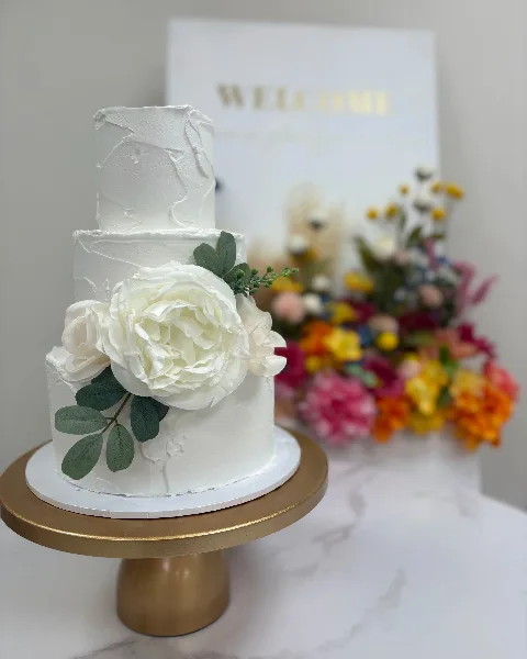 A three-tiered white wedding cake decorated with large white sugar flowers and green leaves, displayed on a gold cake stand.