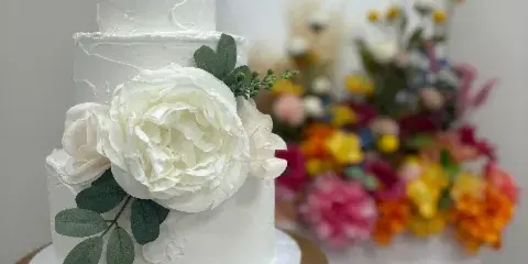 A three-tiered white wedding cake decorated with large white sugar flowers and green leaves, displayed on a gold cake stand.