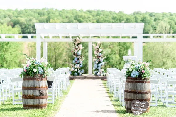 An outdoor wedding ceremony setup featuring a white pergola, rows of white chairs, and floral arrangements in wooden barrels.
