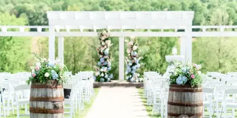 An outdoor wedding ceremony setup featuring a white pergola, rows of white chairs, and floral arrangements in wooden barrels.