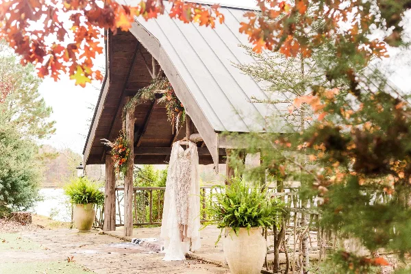 A rustic wooden pavilion with a metal roof, decorated with a hanging wedding dress and floral garlands, situated near a lake with potted ferns in the foreground.