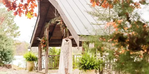A rustic wooden pavilion with a metal roof, decorated with a hanging wedding dress and floral garlands, situated near a lake with potted ferns in the foreground.