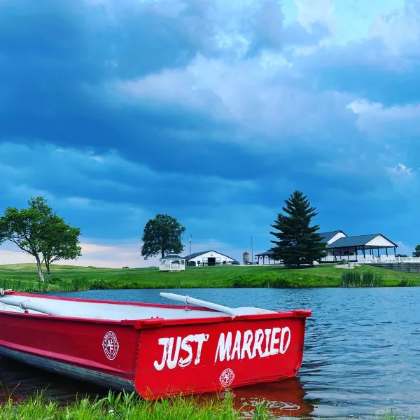 A red boat with 'Just Married' written on the side floats on a lake in front of a white barn and farmhouse on a farm property.