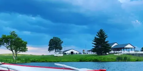 A red boat with 'Just Married' written on the side floats on a lake in front of a white barn and farmhouse on a farm property.