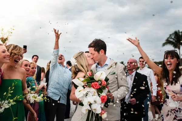 A bride and groom share a kiss while guests throw confetti in the air during an outdoor wedding celebration.