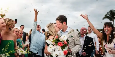 A bride and groom share a kiss while guests throw confetti in the air during an outdoor wedding celebration.