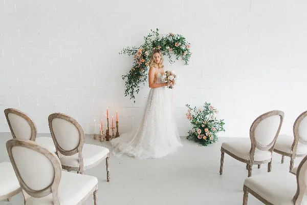 A bride in a white gown holds a bouquet while standing beside a floral arch and floor arrangement in a white room.