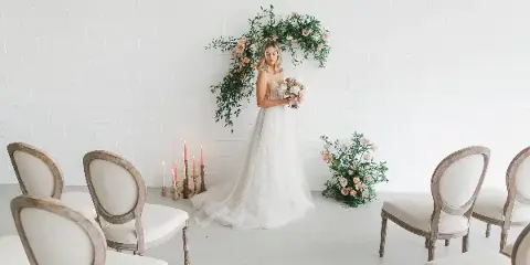 A bride in a white gown holds a bouquet while standing beside a floral arch and floor arrangement in a white room.