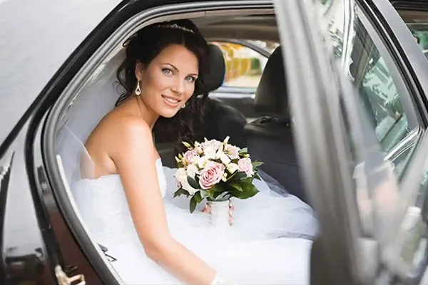 A bride in a white wedding dress and holding a bouquet of flowers seated inside a limousine.