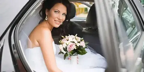 A bride in a white wedding dress and holding a bouquet of flowers seated inside a limousine.