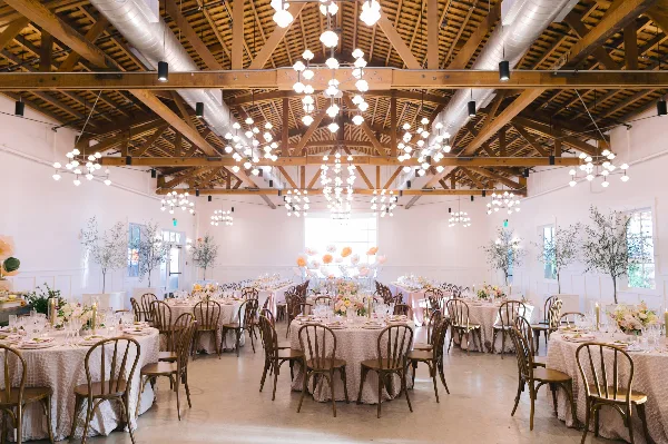 A spacious, elegant banquet hall with wooden beams, modern chandeliers, round tables covered in white linens, wooden chairs, and floral centerpieces set for a wedding or formal event.