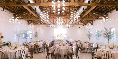 A spacious, elegant banquet hall with wooden beams, modern chandeliers, round tables covered in white linens, wooden chairs, and floral centerpieces set for a wedding or formal event.